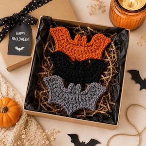 May include: Three crocheted bat ornaments in orange, black, and gray, arranged inside a square cardboard gift box lined with black tissue paper and shredded brown paper filler. A black gift tag attached to the box lid reads HAPPY HALLOWEEN.