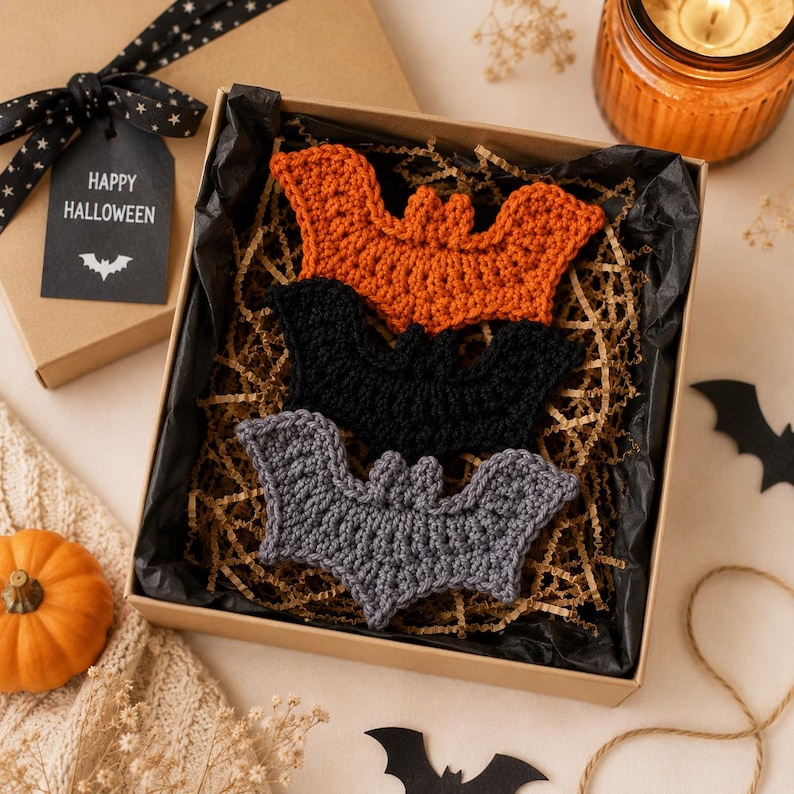 May include: Three crocheted bat ornaments in orange, black, and gray, arranged inside a square cardboard gift box lined with black tissue paper and shredded brown paper filler. A black gift tag attached to the box lid reads HAPPY HALLOWEEN.