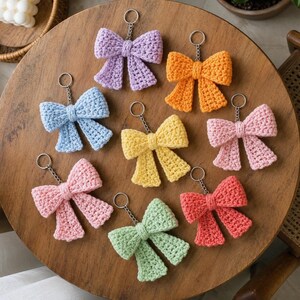May include: Eight crochet bow keychains in various colours including lavender, orange, light blue, yellow, pink, light green, coral, and soft pink. Each bow features a central knot and two hanging tails, attached to a silver metal split ring.