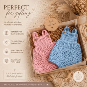 May include: A gift box containing two handmade crochet baby rompers, one in light pink and one in light blue. Each romper features two shoulder straps with small brown buttons. A round cardboard tag labeled Made with love is tied to the box.
