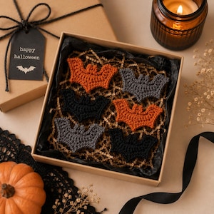 May include: A set of six crocheted bat appliques in orange, black, and gray, arranged inside a square cardboard gift box filled with shredded paper. A black gift tag with the text "happy halloween" and a bat icon is attached to the box lid.