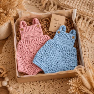 May include: Two crochet baby rompers, one in light pink and one in light blue, are nestled in a cardboard gift box. Each romper features a square neckline, two shoulder straps with small wooden buttons, and a textured stitch pattern throughout.