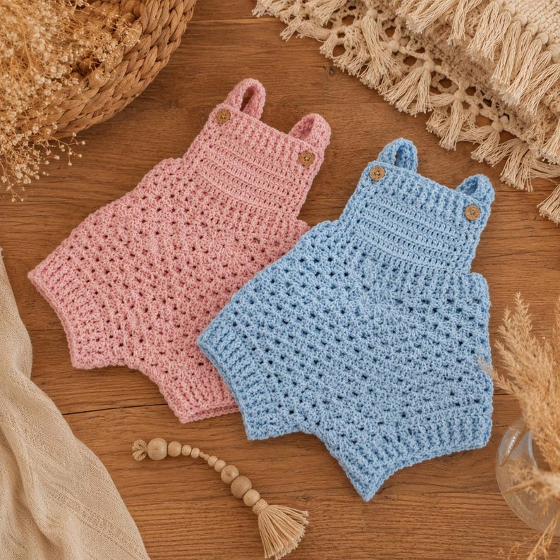 May include: Two crochet rompers, one in light pink and one in light blue, each featuring a square neckline, two shoulder straps with wooden buttons, and a textured stitch pattern. The garments are sleeveless with a short, bloomer-style bottom.