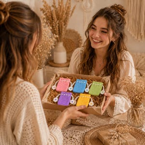 May include: A cardboard gift box containing five small, rectangular, crocheted drawstring pouches in pink, blue, light green, purple, and yellow. Each pouch features a white crocheted flower accent attached to the drawstring cord ends.