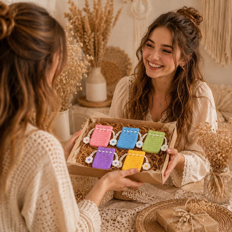 May include: A cardboard gift box containing five small, rectangular, crocheted drawstring pouches in pink, blue, light green, purple, and yellow. Each pouch features a white crocheted flower accent attached to the drawstring cord ends.