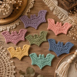 May include: A collection of seven crocheted bat appliques in various colors including purple, peach, dusty pink, olive green, mustard yellow, blue, and mint green, each featuring a distinct wing shape and small pointed ears.