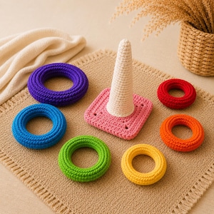 May include: A crochet stacking toy featuring a central off-white cone attached to a square pink base. The set includes five separate circular rings in purple, blue, green, yellow, and red, all crafted with a textured yarn stitch pattern.