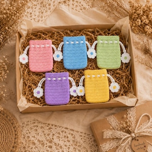 May include: Five small, rectangular, crocheted drawstring pouches in pink, blue, light green, purple, and yellow. Each pouch features a white drawstring with a small, crocheted flower at each end, and a white woven band near the top opening.