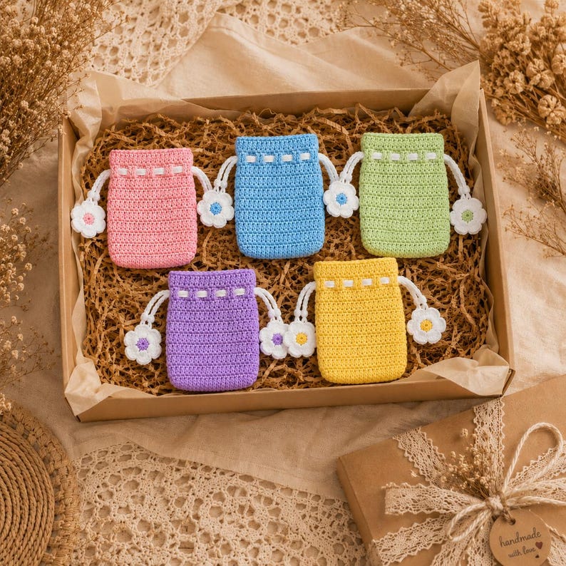 May include: Five small, rectangular, crocheted drawstring pouches in pink, blue, light green, purple, and yellow. Each pouch features a white drawstring with a small, crocheted flower at each end, and a white woven band near the top opening.