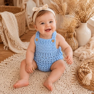 May include: A light blue crocheted romper featuring a textured stitch pattern, two small wooden buttons at the shoulder straps, and a matching cream-colored fabric headband with a bow detail worn on the head.