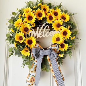 May include: A circular wreath featuring bright yellow sunflowers and green eucalyptus leaves. A wooden cutout script sign reads Welcome in the center. A large decorative bow with a sunflower pattern and navy blue and white stripes hangs below.