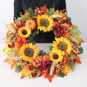 May include: A circular autumn wreath featuring bright yellow sunflowers, orange pumpkins, deep red berries, and brown roses. The arrangement includes various green and orange maple-style leaves, wheat-like stalks, and small clusters of orange berries.