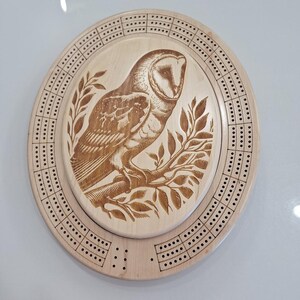 May include: Oval wooden cribbage board featuring a detailed engraving of a barn owl perched on a branch with leaves. The board has a light wood finish and is surrounded by holes for pegging.