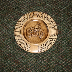 May include: A wooden cribbage board with a carved motorcycle design in the center. The board has a dark brown stain and black painted lines.