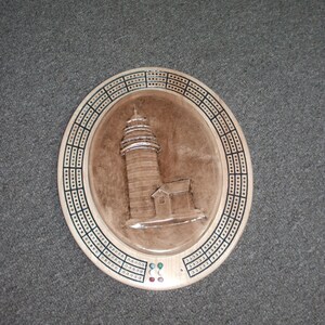 May include: A wooden cribbage board with a lighthouse design. The board has a brown background with a white lighthouse and a small building. The board has a black and white cribbage track around the edge and a red, green, and blue scoring area.