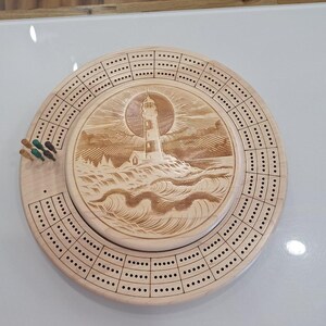 May include: A wooden cribbage board with a detailed lighthouse carving in the center. The board is circular with multiple rows of holes for pegging and includes a set of colored pegs. The design features a lighthouse on a rocky shore.