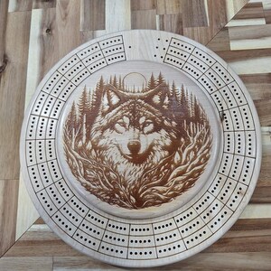 May include: A round wooden cribbage board with a detailed wolf design in the center. The wolf is surrounded by trees and foliage. The board has multiple rows of drilled holes for pegs, and is made of light-colored wood.