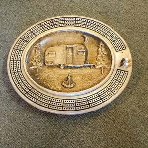 May include: A wooden cribbage board with a carved design of a vintage camper, trees, and a campfire. The board has a black and white checkerboard pattern around the edge.