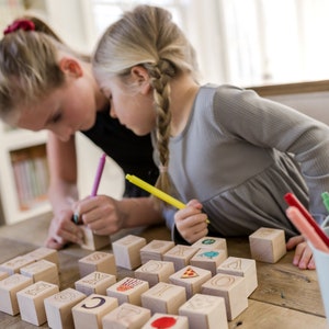 May include: Two young people are focused on wooden blocks with letters and images. They are using colorful markers to draw on the blocks. A white bucket filled with markers sits nearby on a wooden table.
