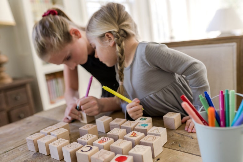 May include: Two young people are focused on wooden blocks with letters and images. They are using colorful markers to draw on the blocks. A white bucket filled with markers sits nearby on a wooden table.