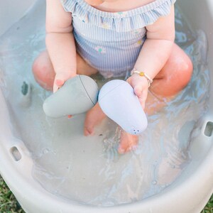 Puede incluir: Un beb&eacute; est&aacute; jugando en una ba&ntilde;era de pl&aacute;stico blanca con dos juguetes de ba&ntilde;o azules y grises con forma de peras. La ba&ntilde;era est&aacute; llena de agua.
