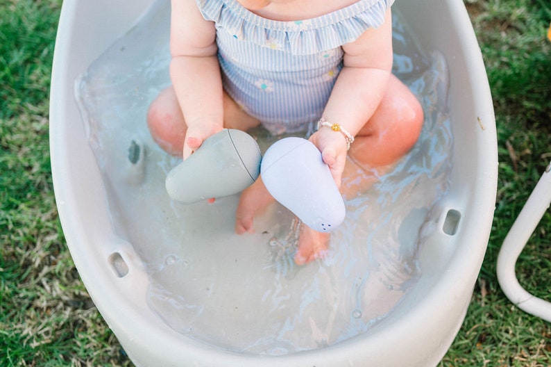 Puede incluir: Un beb&eacute; est&aacute; jugando en una ba&ntilde;era de pl&aacute;stico blanca con dos juguetes de ba&ntilde;o azules y grises con forma de peras. La ba&ntilde;era est&aacute; llena de agua.