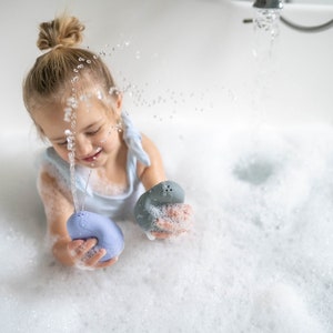 Puede incluir: Una ni&ntilde;a peque&ntilde;a juega en una ba&ntilde;era llena de burbujas. La ni&ntilde;a sostiene dos juguetes de ba&ntilde;o coloridos, uno azul y otro gris. El agua sale a borbotones del grifo.