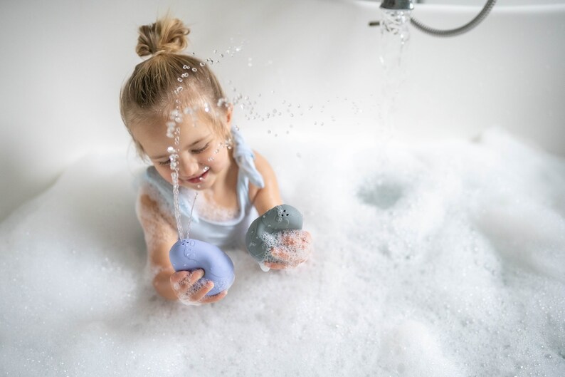 Puede incluir: Una ni&ntilde;a peque&ntilde;a juega en una ba&ntilde;era llena de burbujas. La ni&ntilde;a sostiene dos juguetes de ba&ntilde;o coloridos, uno azul y otro gris. El agua sale a borbotones del grifo.
