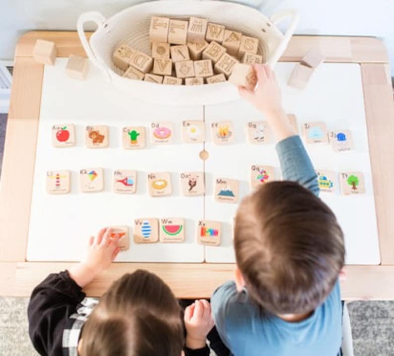 May include: Two children are playing with wooden alphabet blocks. The blocks have letters and pictures on them. The blocks are arranged in a grid on a white table. The children are sitting at the table and are looking at the blocks.