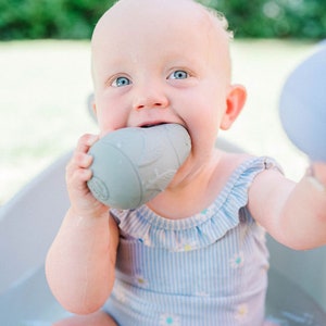 Puede incluir: Un beb&eacute; con un traje de ba&ntilde;o azul y blanco a rayas est&aacute; jugando con dos juguetes de ba&ntilde;o grises y azules en una ba&ntilde;era blanca. El beb&eacute; est&aacute; mordiendo uno de los juguetes y sosteniendo el otro en su mano.