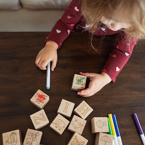 May include: Wooden alphabet blocks with engraved letters and animal illustrations are arranged on a dark wooden surface. A child is holding a block and a marker. The blocks are decorated with drawings in red, green, and other colors. Markers in blue, yellow, and purple are also present.