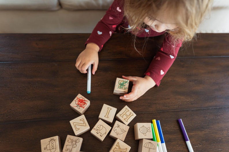 May include: Wooden alphabet blocks with engraved letters and animal illustrations are arranged on a dark wooden surface. A child is holding a block and a marker. The blocks are decorated with drawings in red, green, and other colors. Markers in blue, yellow, and purple are also present.