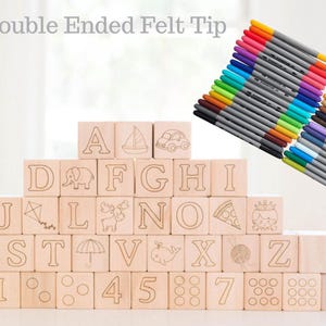 May include: Wooden alphabet blocks with engraved letters and illustrations, stacked in a pyramid shape. A set of double-ended felt tip markers in various colors is in the upper right corner. The text "Double Ended Felt Tip" is at the top.