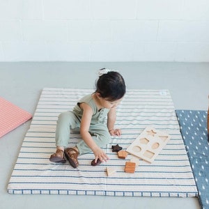 Puede incluir: Un ni&ntilde;o juega con un clasificador de formas de madera y bloques sobre una alfombra de juego a rayas. La alfombra tiene un fondo crema con rayas azul marino. Una alfombra a rayas rosa y una cesta de mimbre tambi&eacute;n est&aacute;n en la imagen.