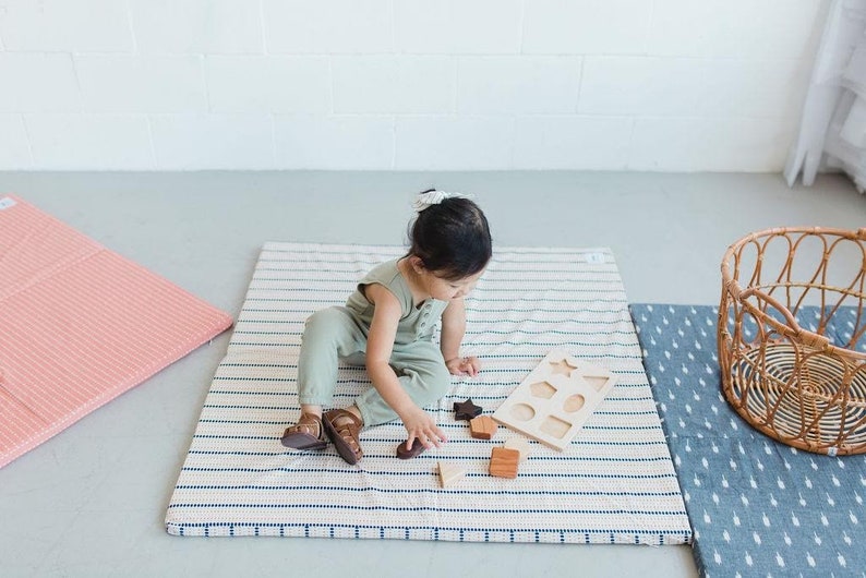 Puede incluir: Un ni&ntilde;o juega con un clasificador de formas de madera y bloques sobre una alfombra de juego a rayas. La alfombra tiene un fondo crema con rayas azul marino. Una alfombra a rayas rosa y una cesta de mimbre tambi&eacute;n est&aacute;n en la imagen.