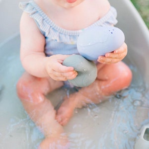 Puede incluir: Un beb&eacute; est&aacute; sentado en una ba&ntilde;era blanca llena de agua, jugando con dos juguetes de ba&ntilde;o suaves y texturizados. Un juguete es azul y el otro es gris.