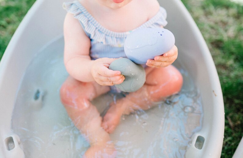 Puede incluir: Un beb&eacute; est&aacute; sentado en una ba&ntilde;era blanca llena de agua, jugando con dos juguetes de ba&ntilde;o suaves y texturizados. Un juguete es azul y el otro es gris.