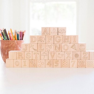 May include: Wooden alphabet blocks with engraved letters, numbers, and symbols are stacked in a pyramid shape. A terracotta pot filled with colored pencils sits to the left. The blocks are on a white surface.
