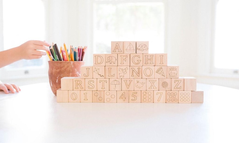 May include: Wooden alphabet blocks with engraved letters, numbers, and symbols are stacked in a pyramid shape. A terracotta pot filled with colored pencils sits to the left. The blocks are on a white surface.