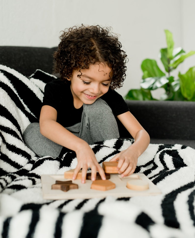 Puede incluir: Un ni&ntilde;o juega con un rompecabezas de formas de madera. El rompecabezas tiene varias formas geom&eacute;tricas en tonos marrones y de madera natural. El ni&ntilde;o lleva una camiseta negra y pantalones grises.