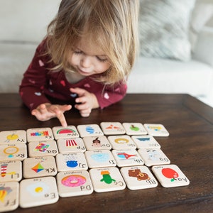 May include: A child plays with a set of wooden alphabet blocks featuring colorful illustrations of various objects. The blocks are arranged in a grid pattern on a wooden table. The letters on the blocks include: Ee, Aa, Bb, C, Dd, 60, Doughnut, LI, Kk, Hh, I, Rr, Qa, a, PP, Oo, Zz, YY, ww.