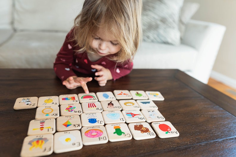 May include: A child plays with a set of wooden alphabet blocks featuring colorful illustrations of various objects. The blocks are arranged in a grid pattern on a wooden table. The letters on the blocks include: Ee, Aa, Bb, C, Dd, 60, Doughnut, LI, Kk, Hh, I, Rr, Qa, a, PP, Oo, Zz, YY, ww.