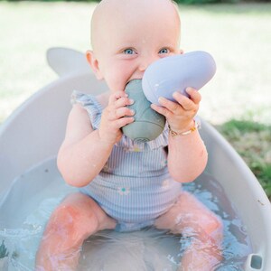 Puede incluir: Un beb&eacute; con un traje de ba&ntilde;o a rayas azul y blanco se sienta en una ba&ntilde;era blanca llena de agua. El beb&eacute; sostiene dos juguetes de ba&ntilde;o, uno morado y otro gris.