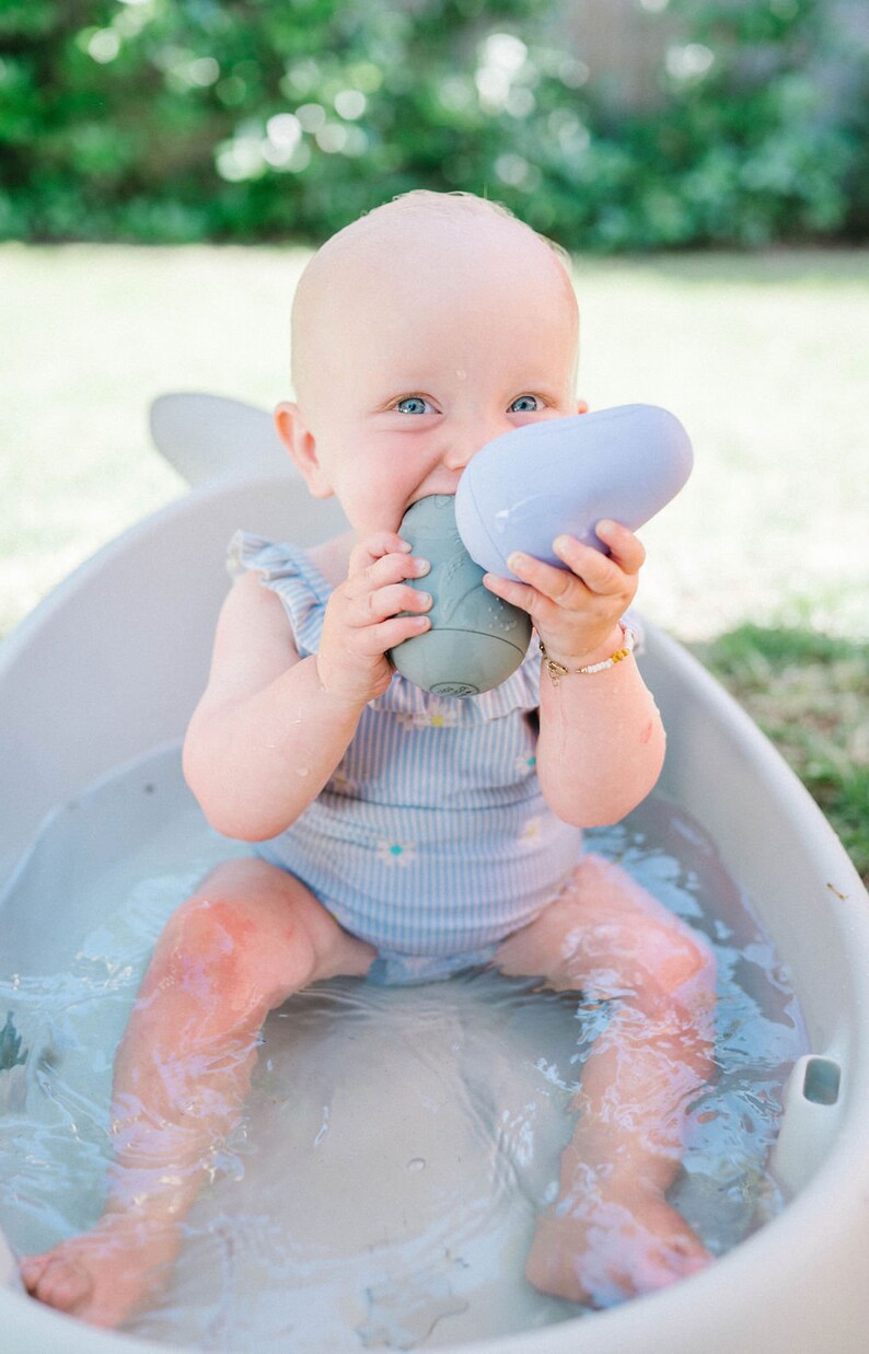 Puede incluir: Un beb&eacute; con un traje de ba&ntilde;o a rayas azul y blanco se sienta en una ba&ntilde;era blanca llena de agua. El beb&eacute; sostiene dos juguetes de ba&ntilde;o, uno morado y otro gris.