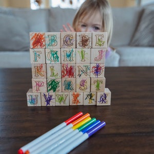 May include: Wooden alphabet blocks with colorful scribbles and a set of rainbow-colored markers on a dark wooden table. A child is partially visible behind the blocks. Educational toy for kids.