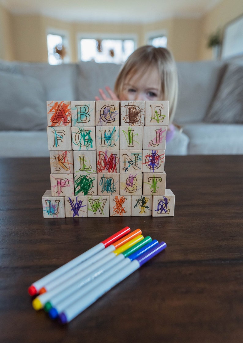 May include: Wooden alphabet blocks with colorful scribbles and a set of rainbow-colored markers on a dark wooden table. A child is partially visible behind the blocks. Educational toy for kids.