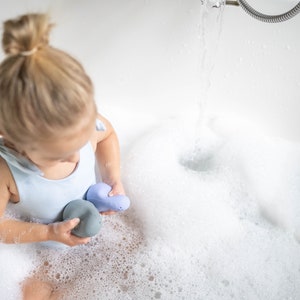 Puede incluir: Una ni&ntilde;a peque&ntilde;a con un traje de ba&ntilde;o azul claro juega con dos juguetes de ba&ntilde;o azules en una ba&ntilde;era blanca llena de burbujas.
