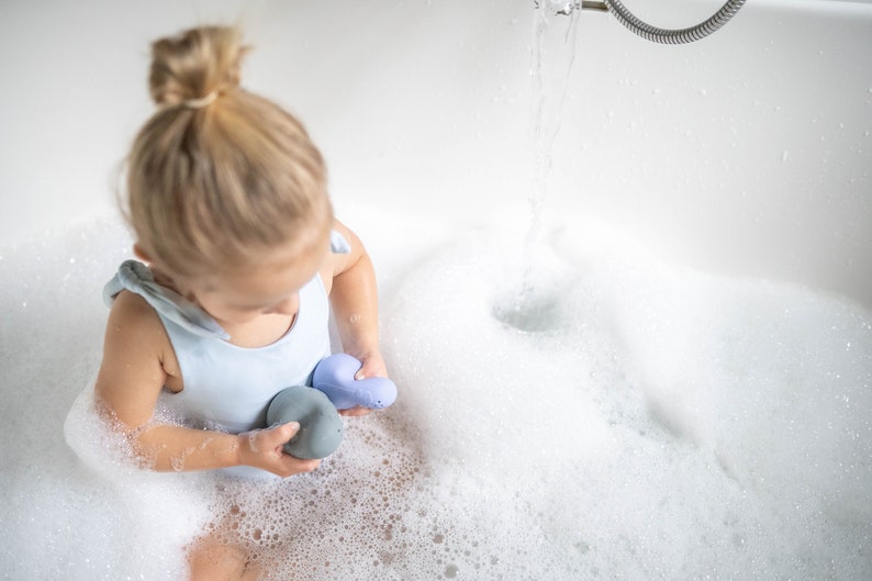 Puede incluir: Una ni&ntilde;a peque&ntilde;a con un traje de ba&ntilde;o azul claro juega con dos juguetes de ba&ntilde;o azules en una ba&ntilde;era blanca llena de burbujas.