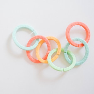 May include: A collection of six interlocking circular rings in pastel shades of mint green, peach, and orange. Each ring features a textured surface with a repeating pattern of small, evenly spaced circular indentations along the outer edge.