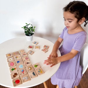 May include: A child sits at a white table playing with wooden alphabet blocks. Each block features a letter of the alphabet and a colorful illustration. The blocks are arranged in alphabetical order.